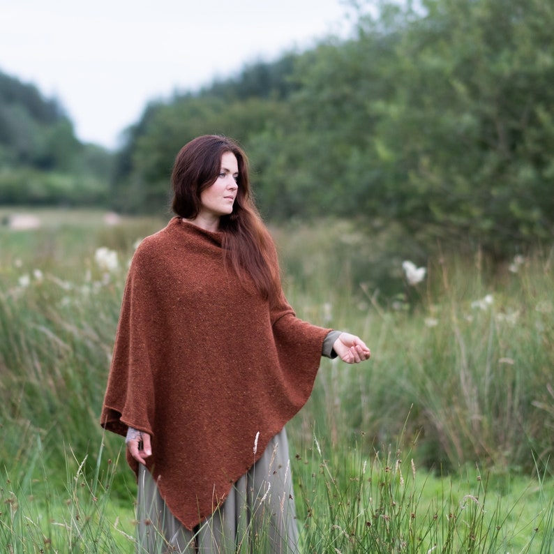 Op de afbeelding: Een vrouw draagt een roestkleurige gebreide poncho in een veld met hoog gras.