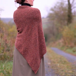 Op de afbeelding: Een roestkleurige gebreide sjaal met een driehoekige vorm, gedragen over een zwarte turtleneck en een bruine rok.