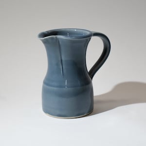 May include: A small, blue ceramic jug with a rounded body and a handle. The jug has a smooth, glossy finish.