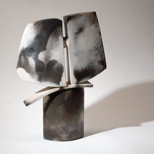 May include: Abstract ceramic sculpture with a white and black glaze. The sculpture is made up of several geometric shapes, including a tall cylinder and two flat, curved pieces.