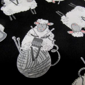 Könnte beinhalten: Ein schwarzer Stoff mit einem sich wiederholenden Muster aus weißen Cartoon-Schafen. Jedes Schaf trägt eine Brille und strickt mit silbernen Nadeln, während es auf einem großen, runden, grauen Wollknäuel sitzt. Die Schafe haben rosa Ohren und Nasen.