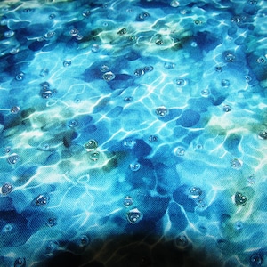 Könnte beinhalten: Nahaufnahme eines Stoffes mit einem wasserähnlichen Design. Das Muster zeigt Blau- und Grüntöne mit weißen Highlights, die Sonnenlicht auf dem Wasser imitieren. Kleine, klare Blasen sind über die Oberfläche verstreut und verstärken das Wasser-Thema.