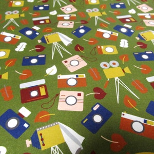 Op de afbeelding: Een groene stof met een herhalend patroon van camera's en filmcamera's. De camera's zijn in verschillende kleuren, waaronder rood, blauw, geel en roze.