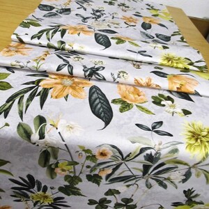 Könnte beinhalten: Ein floraler Tischläufer mit hellgrauem Hintergrund und einem Muster aus orangefarbenen und gelben Blumen, grünen Blättern und weißen Blüten. Der Stoff ist gefaltet und zeigt das volle Design. Der Läufer liegt auf einer hellbraunen Oberfläche.