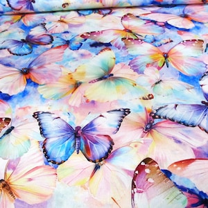Può includere: Un tessuto con un design ad acquerello con numerose farfalle nei toni del blu, rosa, giallo e viola. Le farfalle hanno motivi alari dettagliati e sono sparse su uno sfondo azzurro.