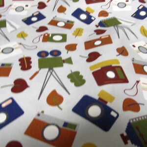 Peut inclure: Un tissu blanc avec un motif répété de caméras, de caméras argentiques et de feuilles dans des tons de rouge, orange, jaune, vert, bleu et marron.