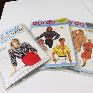 Peut inclure: Trois paquets de patrons de couture vintage. Un paquet est étiqueté "neue mode" avec une femme en chemisier zébré et jupe rouge. Deux paquets sont étiquetés "Burda" avec des illustrations de chemisiers et robes pour femmes.