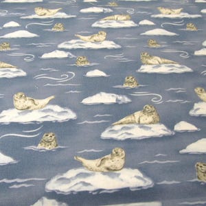 Peut inclure: Un tissu bleu avec un motif répétitif de phoques de couleur claire se reposant sur des banquises blanches. Le design comprend de petites lignes blanches tourbillonnantes représentant le vent ou le mouvement de l'eau sur le fond bleu.
