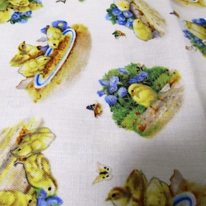 Op de afbeelding: Witte stof met een herhalend patroon van gele kuikens en blauwe bloemen. De kuikens zijn afgebeeld in verschillende poses, sommige in nesten, sommige in groepen en sommige met eieren. De bloemen zijn blauw en wit en hebben een delicate, aquarelachtige uitstraling.