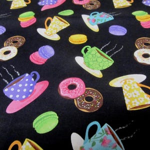 Könnte beinhalten: Schwarzer Stoff mit einem sich wiederholenden Muster aus bunten Teetassen, Untertassen, Donuts und Macarons. Die Teetassen haben Tupfen und florale Designs. Donuts sind rosa und schokoladenfarben, und Macarons sind rosa, grün und lila.