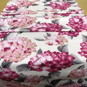May include: A floral table runner featuring a pattern of pink and magenta hydrangea blooms on a cream background. The runner is folded, showcasing the fabric's design and texture. Gray leaves accent the flowers, creating a vibrant, decorative piece.