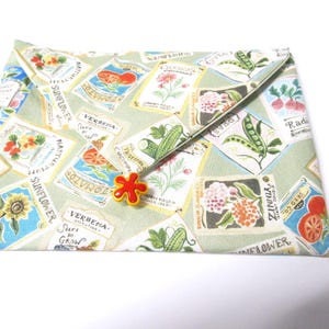 Könnte beinhalten: Eine hellgrüne Stofftasche mit Klappenverschluss, verziert mit Vintage-Saatpäckchen-Designs. Die Tasche wird mit einem rot-gelben blütenförmigen Knopf verschlossen. Die Saatpäckchen zeigen Illustrationen von Sonnenblumen, Tomaten und anderem Gemüse.