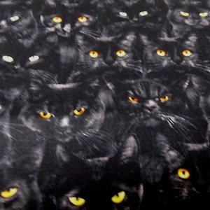 Op de afbeelding: Een stofpatroon met een dichte opstelling van zwarte kattengezichten. Elke kat heeft heldergele of witte ogen, wat een opvallend contrast vormt met de donkere vacht. De gezichtsuitdrukkingen van de katten variëren.