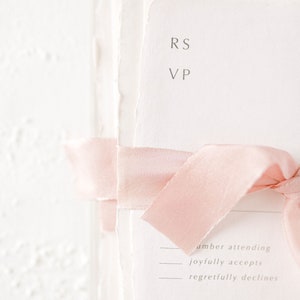 May include: A white RSVP card with a pink ribbon tied around it. The card has the initials "RS" and "VP" printed on it. The card also has the text "number attending", "joyfully accepts", and "regretfully declines" printed on it.