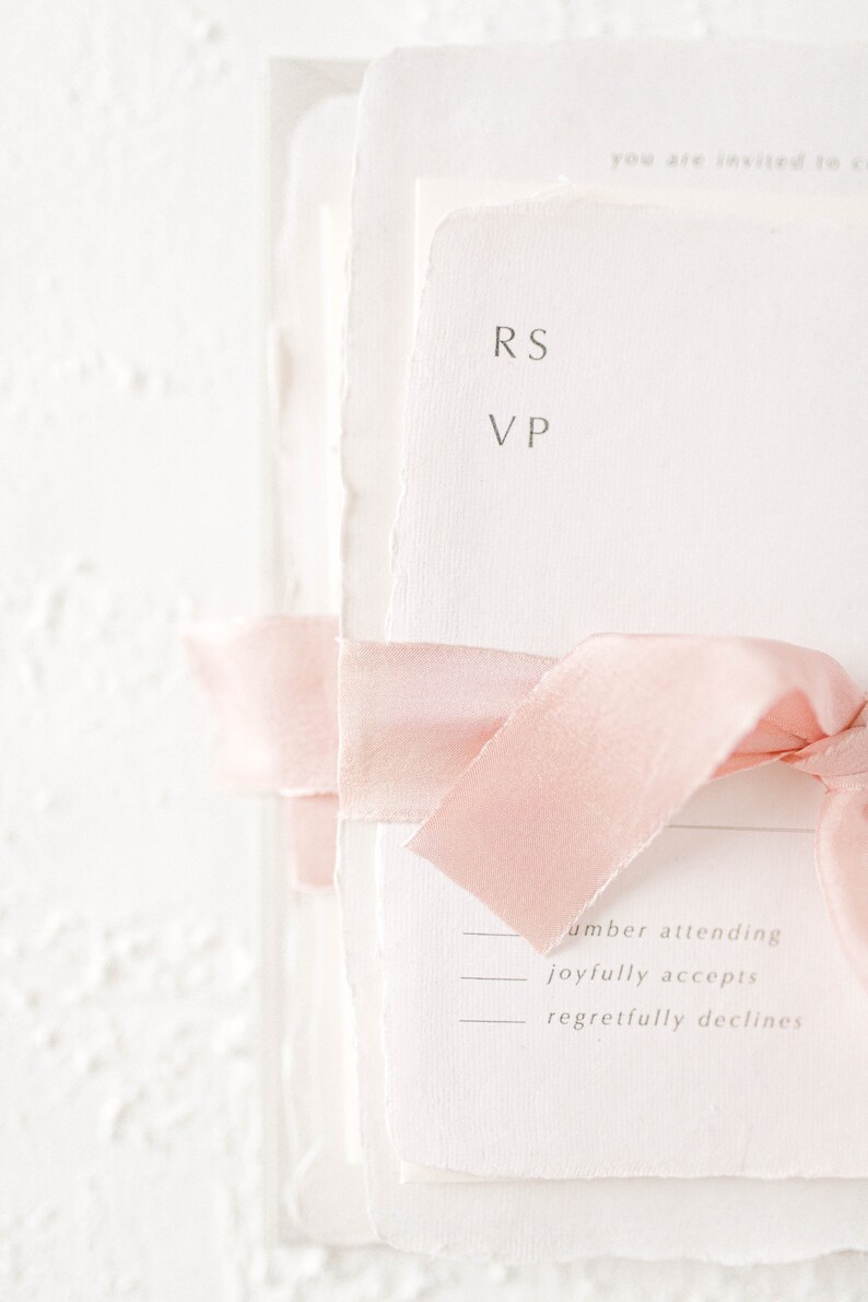 May include: A white RSVP card with a pink ribbon tied around it. The card has the initials "RS" and "VP" printed on it. The card also has the text "number attending", "joyfully accepts", and "regretfully declines" printed on it.