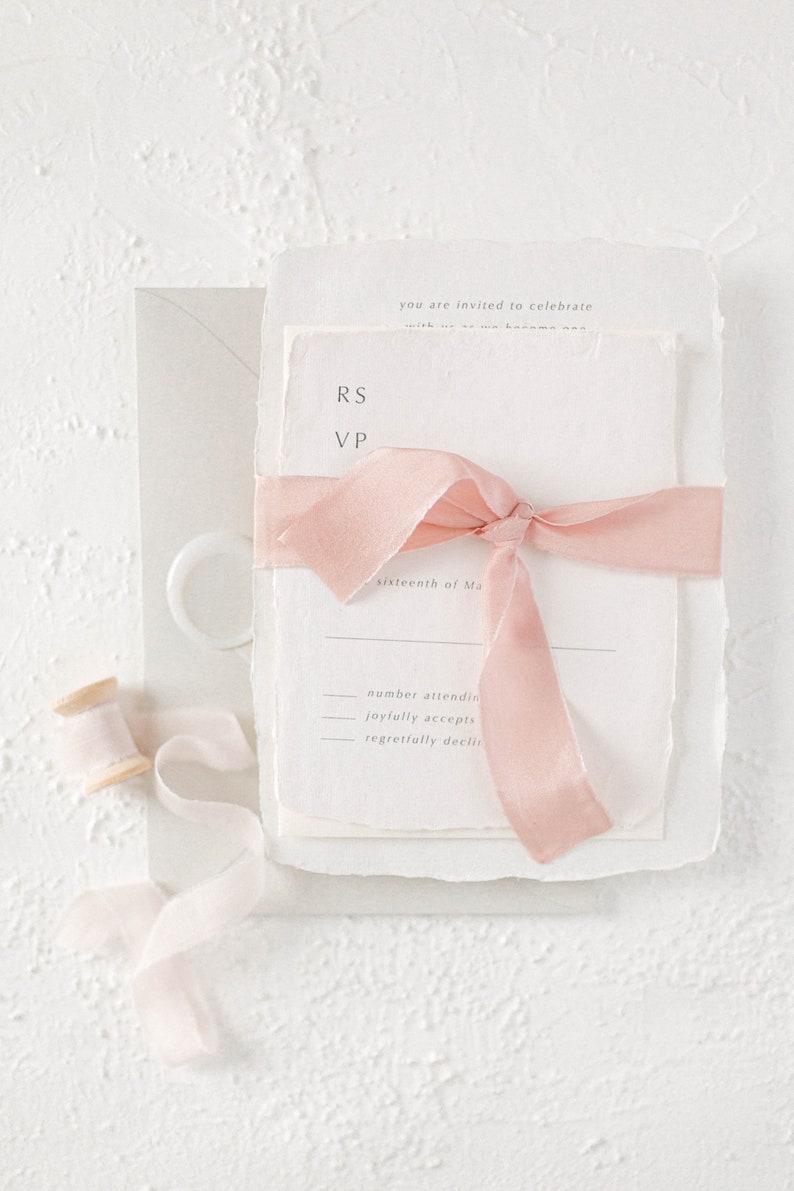 May include: A white wedding invitation with a pink ribbon tied around it. The invitation has the text "RS VP" and "sixteenth of Ma" printed on it. The invitation also has the text "number attending", "joyfully accepts", and "regretfully declines" printed on it.