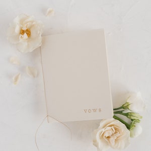 Op de afbeelding: Een wit boek met het woord "VOWS" gedrukt op de voorkant. Het boek is vastgebonden met een dun bruin touwtje. Witte bloemen zijn rondom het boek verspreid.