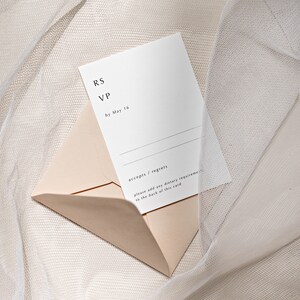 May include: A white RSVP card with black text that reads "RS VP by May 16". The card is partially inside a light pink envelope. The envelope and card are on a white, textured surface.