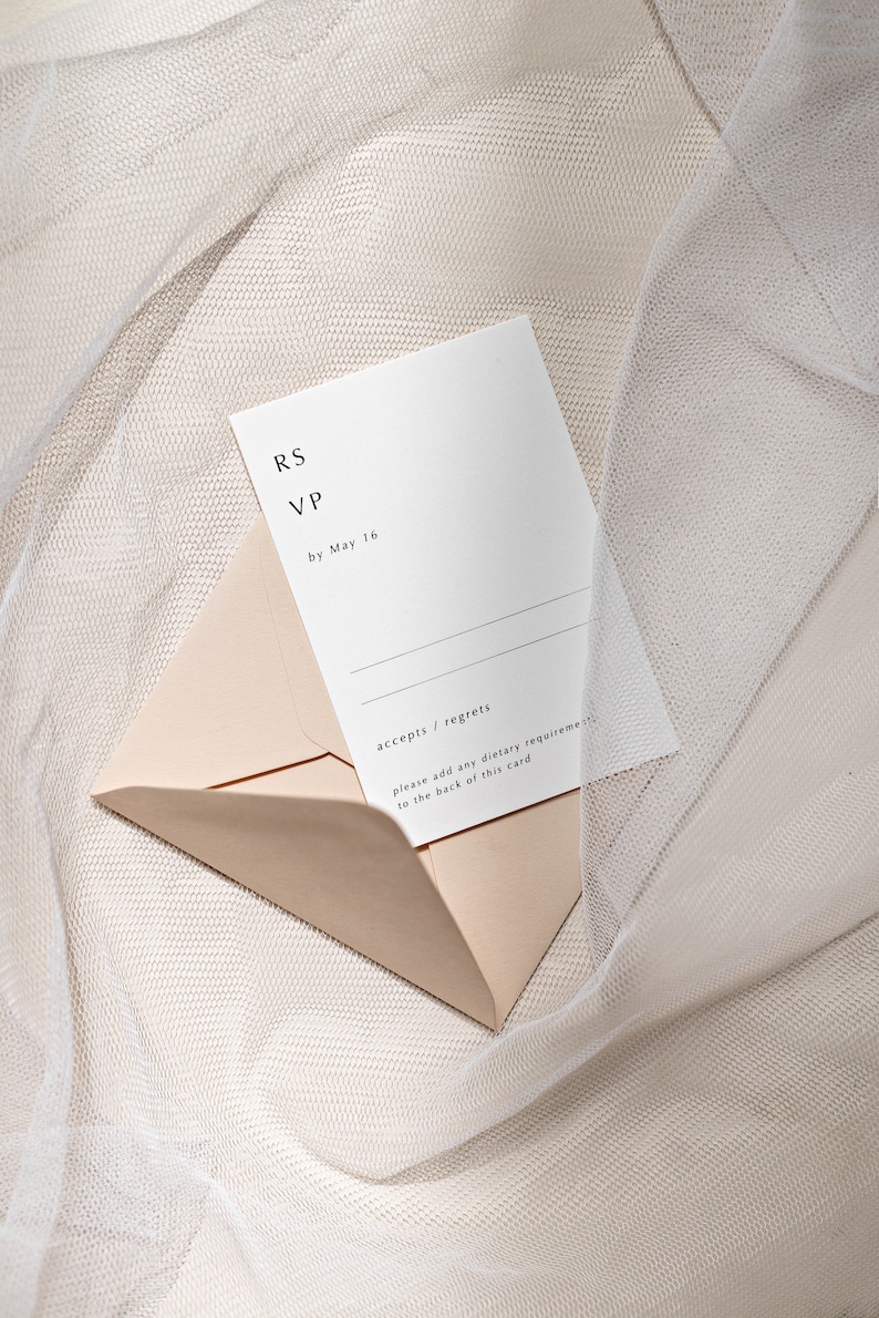 May include: A white RSVP card with black text that reads "RS VP by May 16". The card is partially inside a light pink envelope. The envelope and card are on a white, textured surface.
