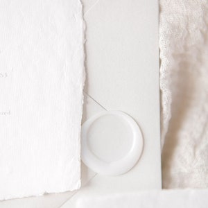 May include: A white envelope with a white wax seal. The envelope is on a white fabric surface.