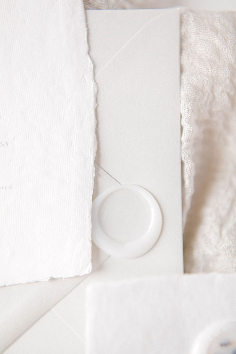 May include: A white envelope with a white wax seal. The envelope is on a white fabric surface.
