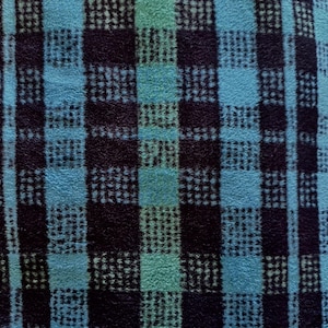 May include: Close-up of a textured fabric with a plaid pattern. The design features squares and stripes in shades of teal, turquoise, and navy blue. The fabric appears soft and plush, with a slightly raised texture.