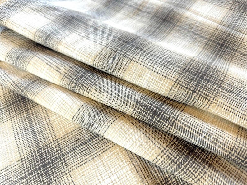 May include: Close-up of a beige and gray plaid fabric with a textured weave. The fabric is folded in a few places, showing the pattern and texture.