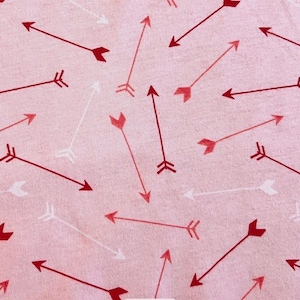 May include: A pink fabric with a repeating pattern of arrows in shades of red and white. The arrows are of varying sizes and directions, creating a scattered design. The fabric has a soft, textured appearance.