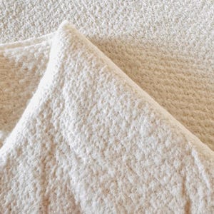 May include: Close-up of a folded, off-white fabric with a textured, nubby surface. The fabric is folded over, creating a triangular shape. The material appears to be thick and possibly absorbent, suitable for towels or blankets.