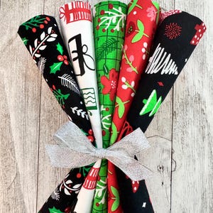 May include: A bundle of six rolled fabric pieces tied with a silver ribbon. The fabrics feature Christmas-themed patterns in red, green, black, and white. Designs include holly, gifts, and Christmas trees.