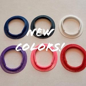 Op de afbeelding: Een assortiment van zes ovale objecten in verschillende kleuren: marineblauw, zwart, wit, paars, roze en rood. De tekst "NEW COLORS!" staat eroverheen. De objecten hebben een textuur.