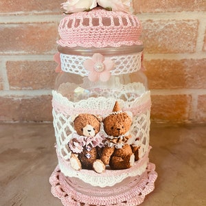 Peut inclure: Un bocal en verre transparent avec un couvercle et une base en crochet rose, orné de dentelle blanche et d'accents floraux roses. Deux ours en peluche sont représentés sur le bocal. Le bocal est décoré de fleurs artificielles sur le dessus.