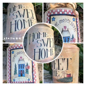 Könnte beinhalten: Eine dekorative Dose mit einem abgenutzten, cremefarbenen Finish. Die Dose hat einen rot-weiß-blauen karierten Rand und den Text "Home Sweet Home" in schwarzen Buchstaben. Die Dose hat auch ein kleines, bemaltes Haus mit einem roten Herzen und den Text "America" darunter.