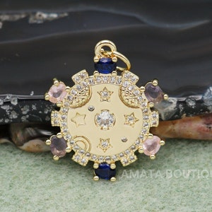 May include: Gold-toned charm with a celestial design featuring a crescent moon, stars, and small pink and blue crystals. The charm has a textured, circular shape and a loop for attaching to a necklace or bracelet.