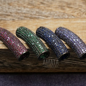 May include: Four curved, cylindrical jewelry components in shades of red, green, blue, and purple, each encrusted with small, sparkling stones. The components are arranged on a wooden surface, with the text "AMATA BOUTIQUE" visible.