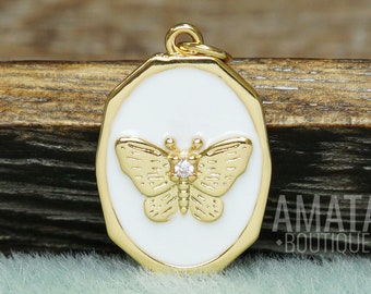 May include: An oval gold-toned pendant featuring a white enamel inlay and a central gold butterfly motif with a small clear stone at its center. The pendant includes a small top loop for attachment and displays the text AMATA BOUTIQUE.