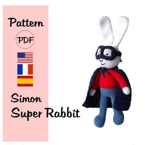 Könnte beinhalten: Eine gehäkelte Simon Super Rabbit Superheldenpuppe mit marineblauer Kapuze und Maske, rotem Hemd und blauer Hose. Das Bild enthält ein rosa Banner mit dem Text "Pattern PDF" und den Flaggen der USA, Frankreichs und Spaniens.