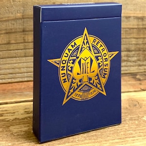 May include: A navy blue deck of playing cards with a gold emblem featuring a spade, star, and the words "NUNQUAM RETRORSUM" and the numbers "1776 1876". The card box is rectangular and stands upright.