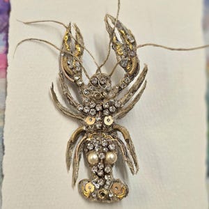 Könnte beinhalten: Aufwendig gearbeitete Hummer-Brosche. Der Hummer besteht aus silberfarbenem Metall, verziert mit klaren Strasssteinen, goldenen Pailletten und Kunstperlen. Die Scheren und Antennen des Hummers sind ausladend.