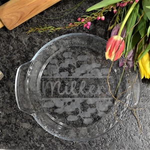 May include: A clear glass pie plate with a floral design and the word "Miller" etched into the bottom. The plate is on a dark gray surface with a rolling pin, a measuring cup of flour, and a bouquet of tulips.