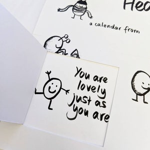 May include: A white greeting card with a cut-out window, featuring a black and white illustration of a smiling character with the text "You are lovely just as you are." Other whimsical drawings and text are visible.