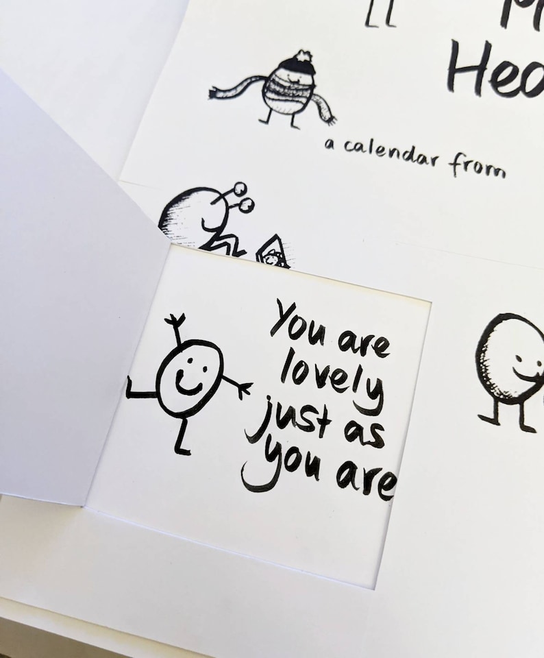 May include: A white greeting card with a cut-out window, featuring a black and white illustration of a smiling character with the text "You are lovely just as you are." Other whimsical drawings and text are visible.