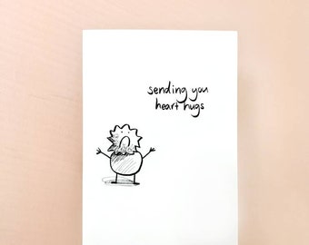 Könnte beinhalten: Eine weiße Grußkarte mit dem handgeschriebenen Text "sending you heart hugs". Unter dem Text befindet sich eine kleine, skurrile Schwarz-Weiß-Illustration einer Kreatur mit ausgestreckten Armen. Die Karte liegt auf einer hellrosa Oberfläche.