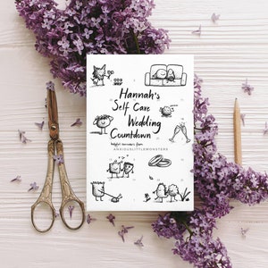 May include: A white card titled "Hannah's Self Care Wedding Countdown" with whimsical illustrations, surrounded by purple lilac flowers, vintage scissors, and a pencil on a white wooden surface.
