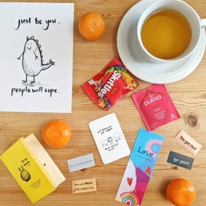 May include: A collection of items on a wooden surface, including a drawing with the text "just be you. people will cope.", a cup of tea, tangerines, sweets, tea bags, and cards with positive messages such as "You are fabulous".