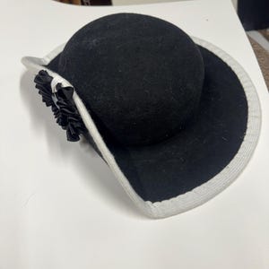 Może przedstawiać: Czarny kapelusz z zaokrągloną koroną i szerokim rondem. Rondo ma jasnoszarą obwódkę. Czarny kwiat z wstążki jest przymocowany z boku. Stylowy dodatek.