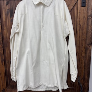 May include: Cream-colored long-sleeve shirt with a collared neckline and a button-down placket. The shirt has a relaxed fit and is made of a textured fabric. The shirt is hanging on a wooden hanger against a dark wooden background.
