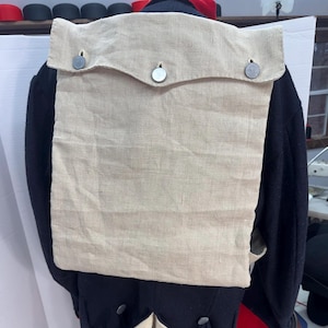 May include: A historical military uniform jacket, primarily black with red trim. A tan-colored linen flap is attached to the back with three silver-colored buttons. The jacket has decorative buttons and a tailored fit, suggesting a formal or ceremonial garment.
