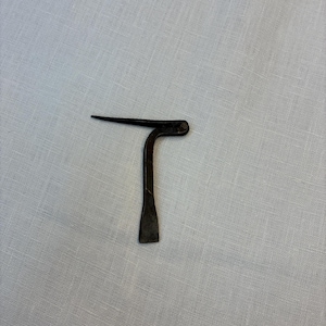 May include: A small, antique metal tool with a dark, aged appearance. The tool has a flat, rectangular base, a curved midsection, and a pointed tip. The overall shape resembles a stylized letter "T".