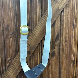 May include: A pale blue leather shoulder strap with a gold-coloured buckle and a dark blue leather accent. The strap is hanging against a wooden background.